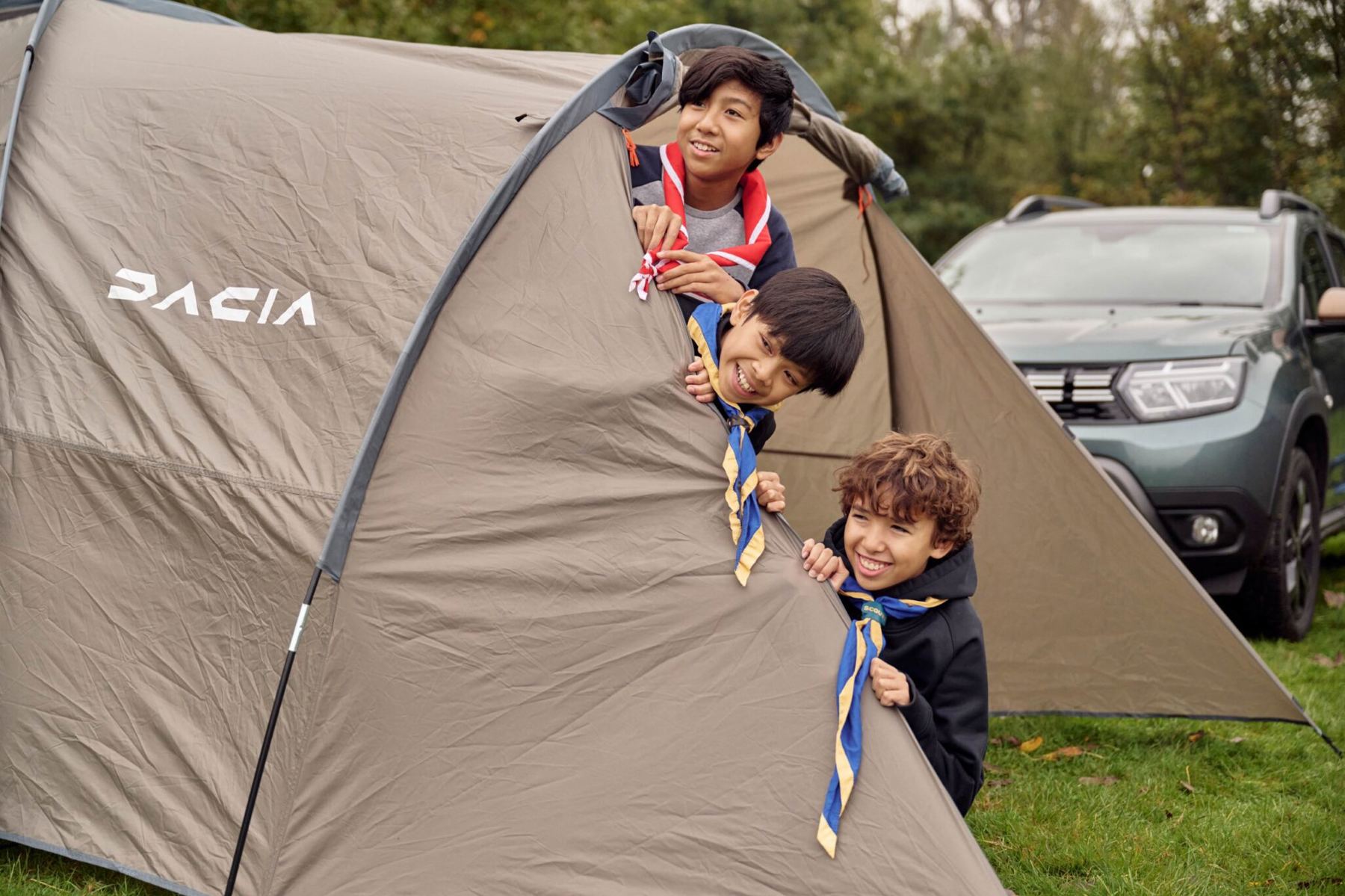 The image shows three Scouts peering out of a beige tent with the Dacia logo in white on the slide. The camera is facing the tent side on, with the three Scouts smiling and wearing neckers. There's a Dacia car just behind the tent.