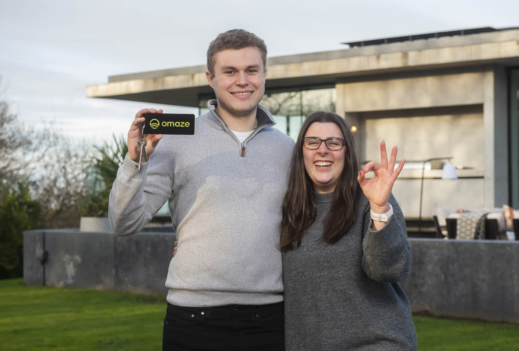 The image shows the winners of the Omaze Million Pound House Draw. They're standing outside in front of their new home in the Wirral, with Ryan to the left holding a key with 'Omaze' written on it, and Lauren to the left holding her hand up with her fingers doing the 'OK' symbol.