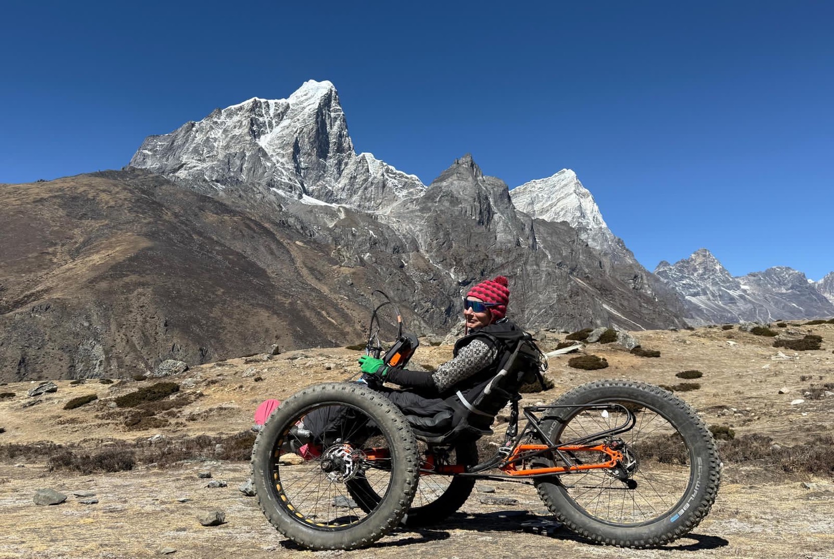 The image shows Karen sitting on her bike on top of a rocky terrain, with mountains and blue sky in the background. She's wearing a red beanie hat, green gloves and sunglasses.
