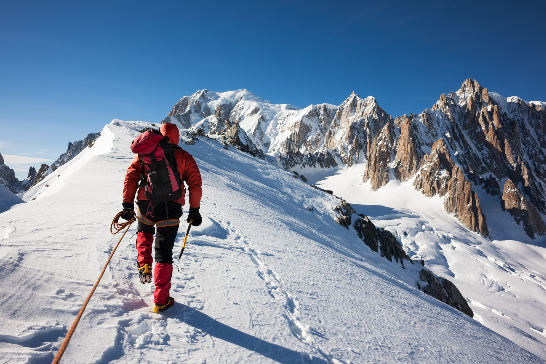 Darren is walking along a snowy mountainside. The camera is behind him as he walks uphill. There are other mountains to the right of him and the sky is bright blue.