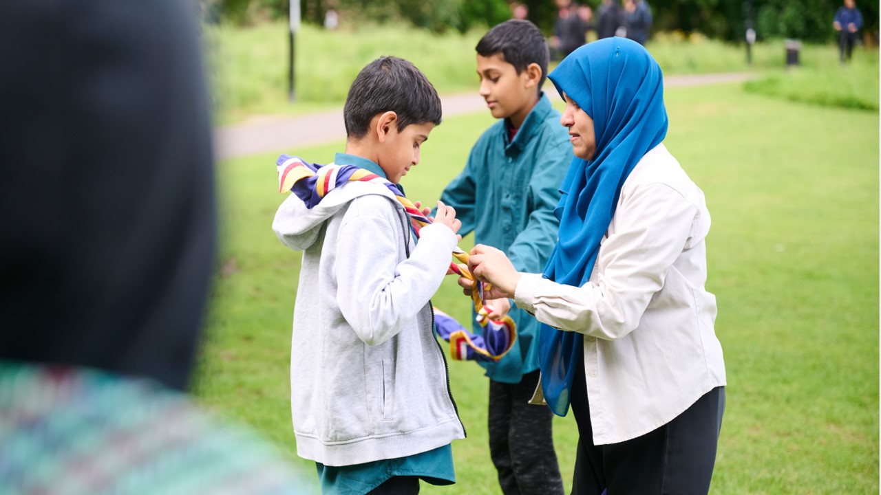 A volunteer, wearing a hijab, ties the necker of a young person wearing a grey hoodie.