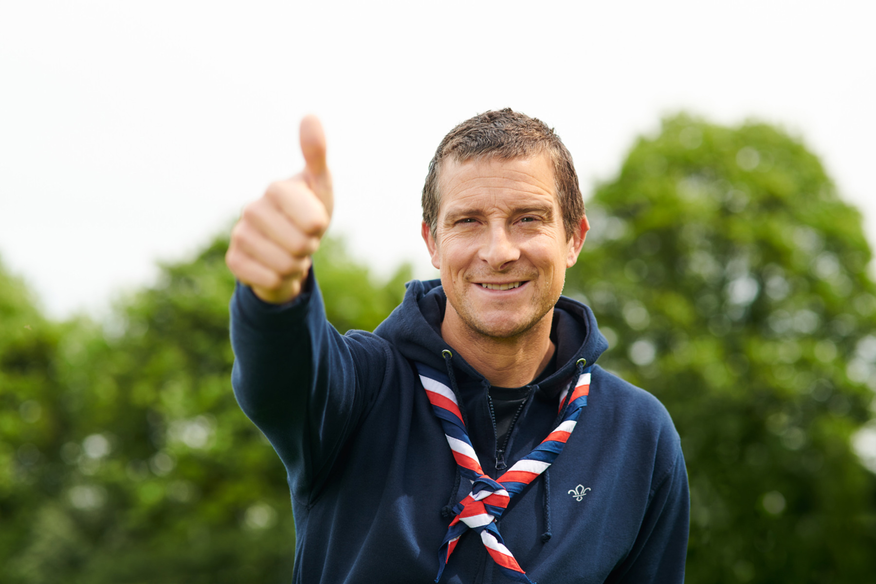 Bear Grylls gives a thumbs up to camera wearing Scouts hoodie and stripy necker scarf with trees in background