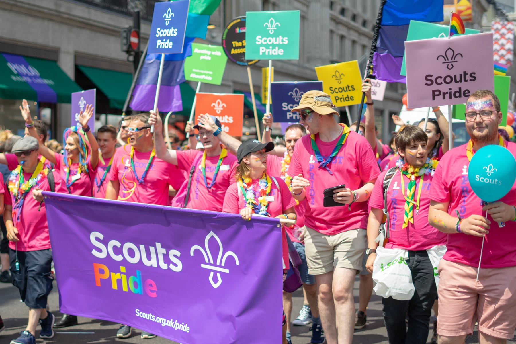 A group of Scouts wearing pink shirts and pride neckers walk in a Pride parade holding a 'Scout Pride' banner.