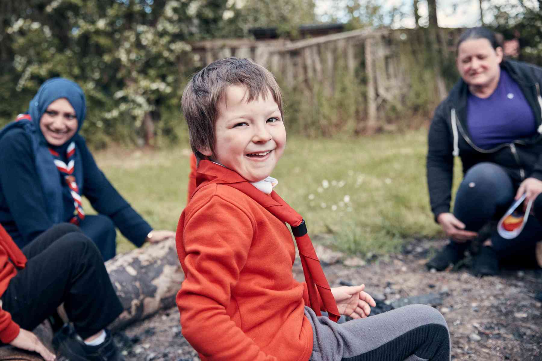 A young person, wearing a red jumper, smiles at the camera while two volunteers sit behind