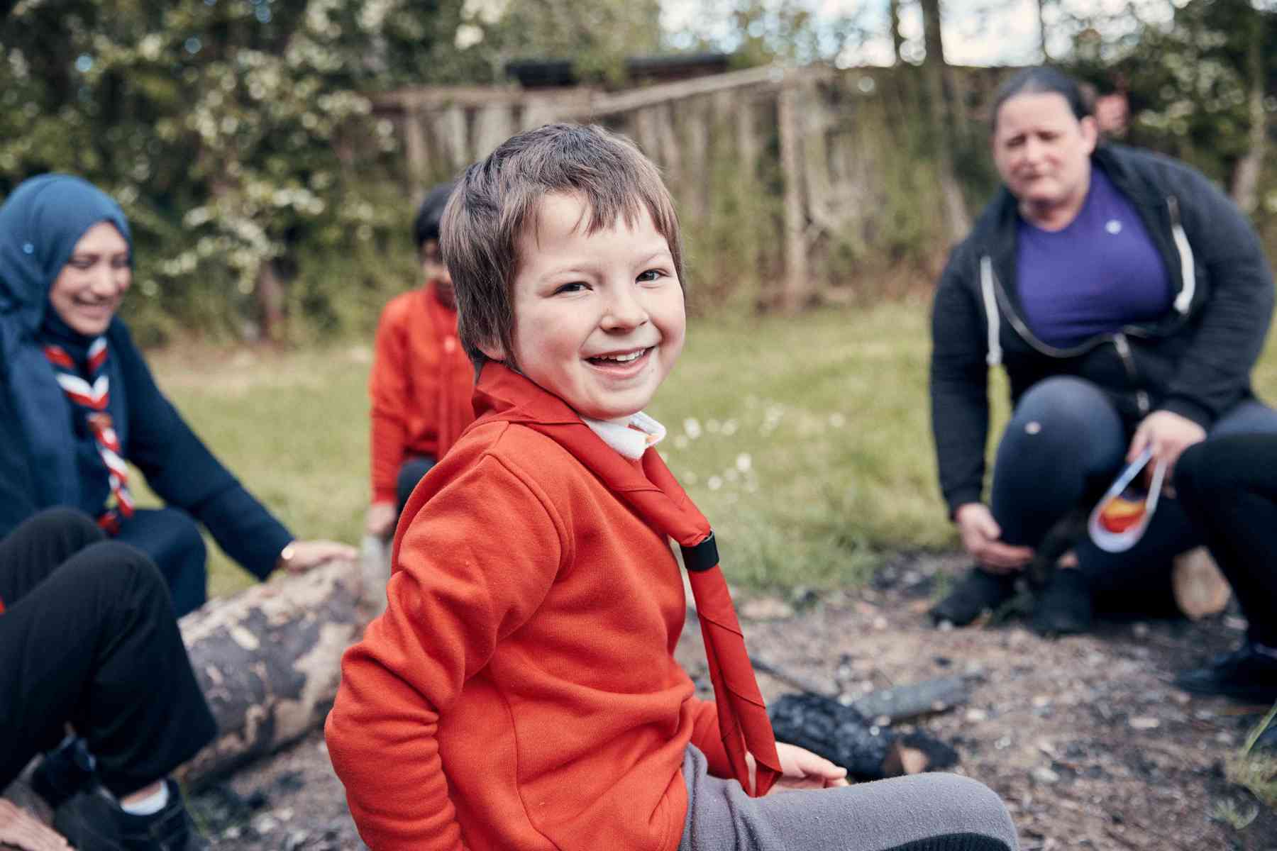 A young person, wearing a red jumper, smiles at the camera while two volunteers sit behind