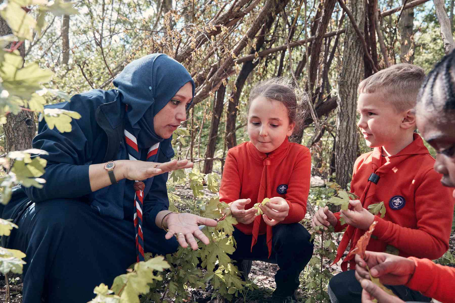 A leader crouches down to speak to three Squirrels, wearing red jumpers and scarves, in the woods.