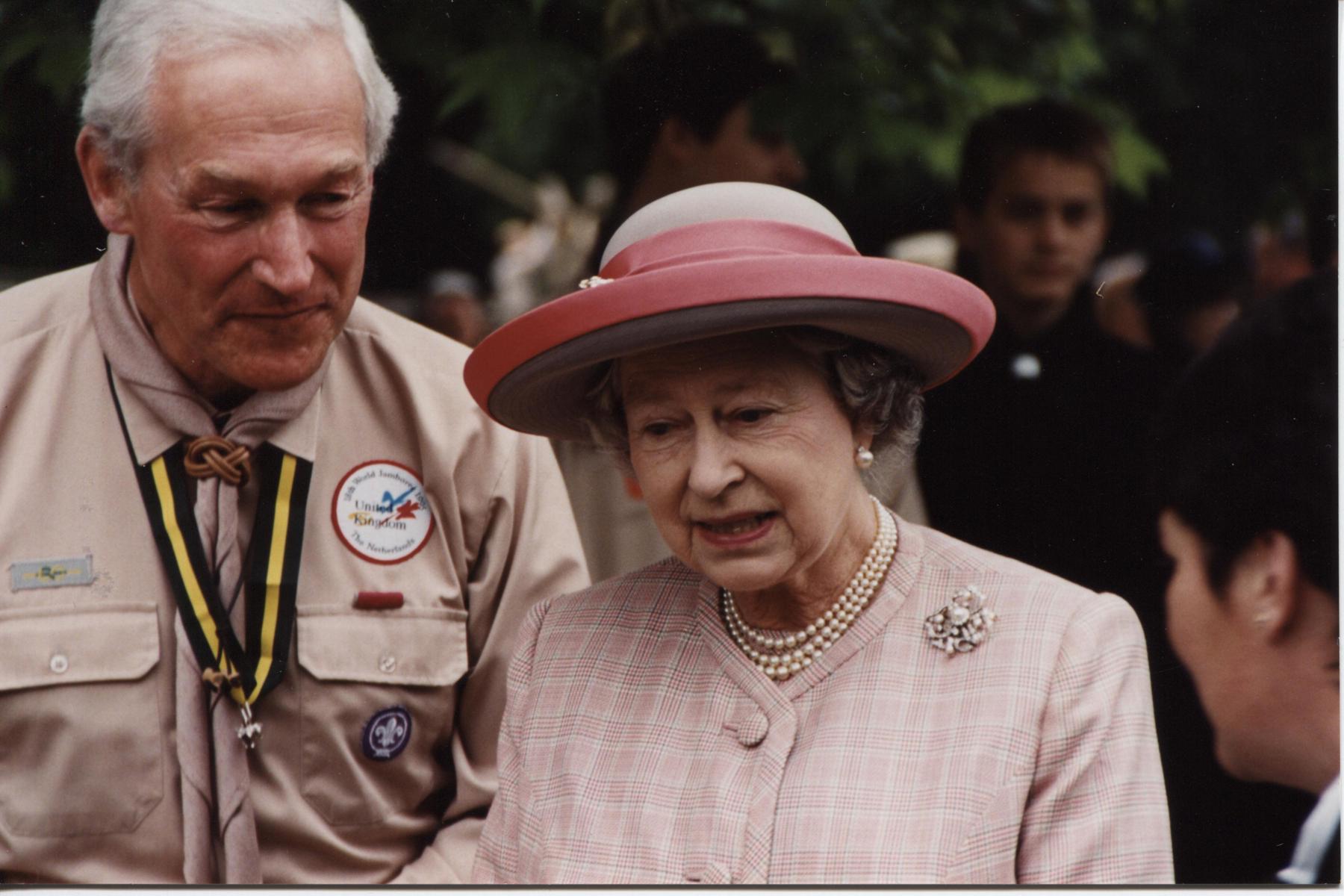 The Queen, wearing a pink hat, talks to a volunteer next to her.