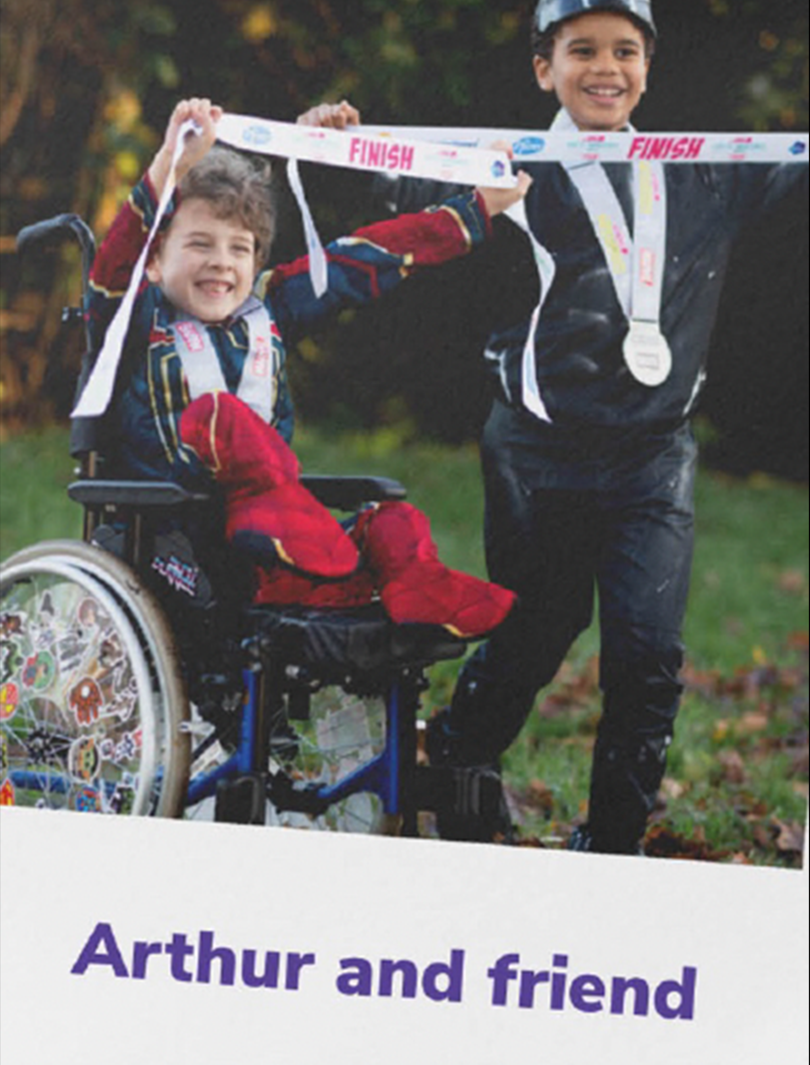 Arthur, in a wheelchair, smiles while holding up a 'finish' banner with his friend.