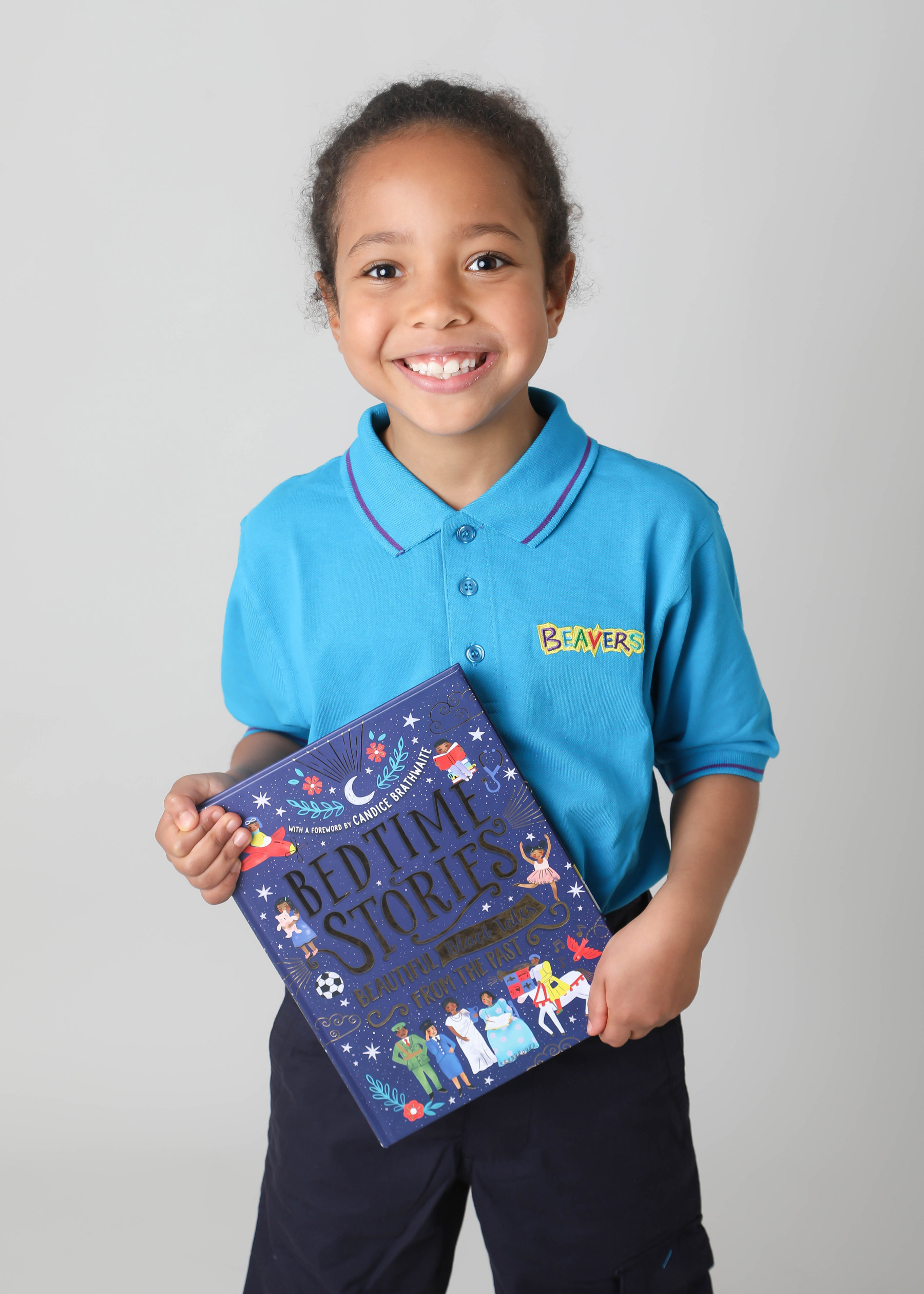 Sarah Mirkin smiling at the camera while wearing a blue Beavers polo shirt and holding a book titled 'Bedtime Stories'