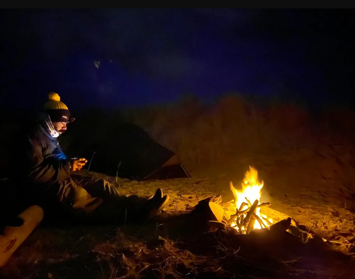 Chris the coast walker sat in a hat by a fire at night time