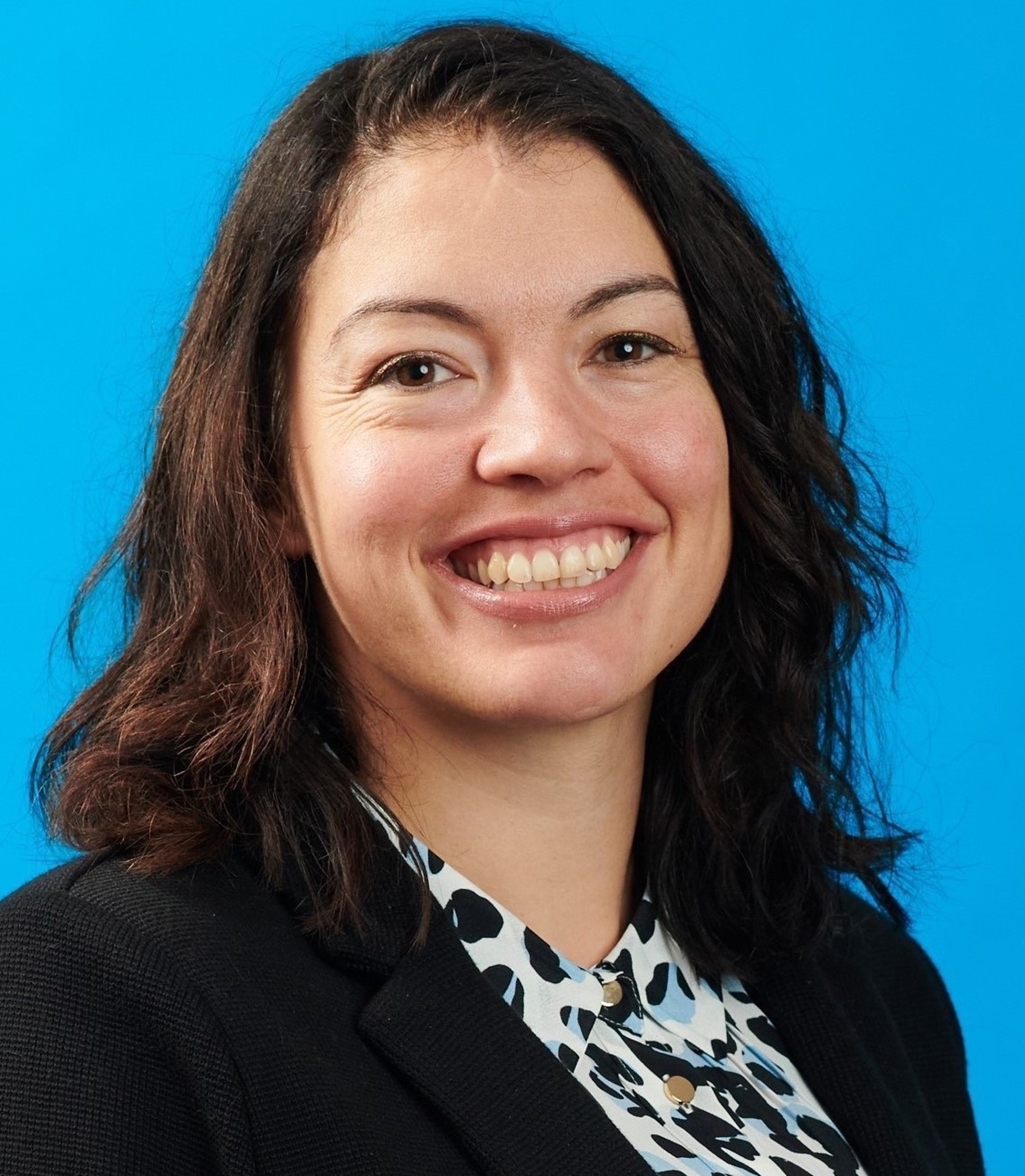 Headshot of Chloe Kembery smiling at the camera wearing a black, white and blue patterned shirt and black blazer