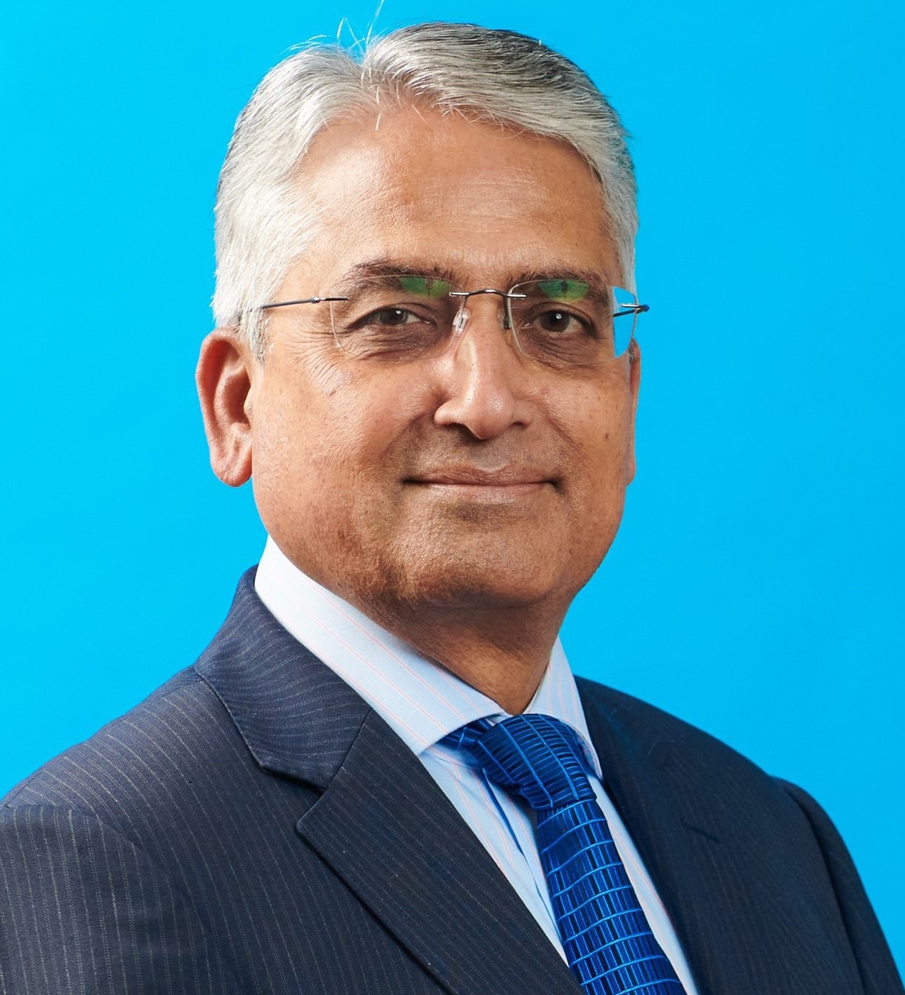 Headshot of Kris Murali smiling at the camera wearing glasses, a white shirt, blue tie and navy suit jacket