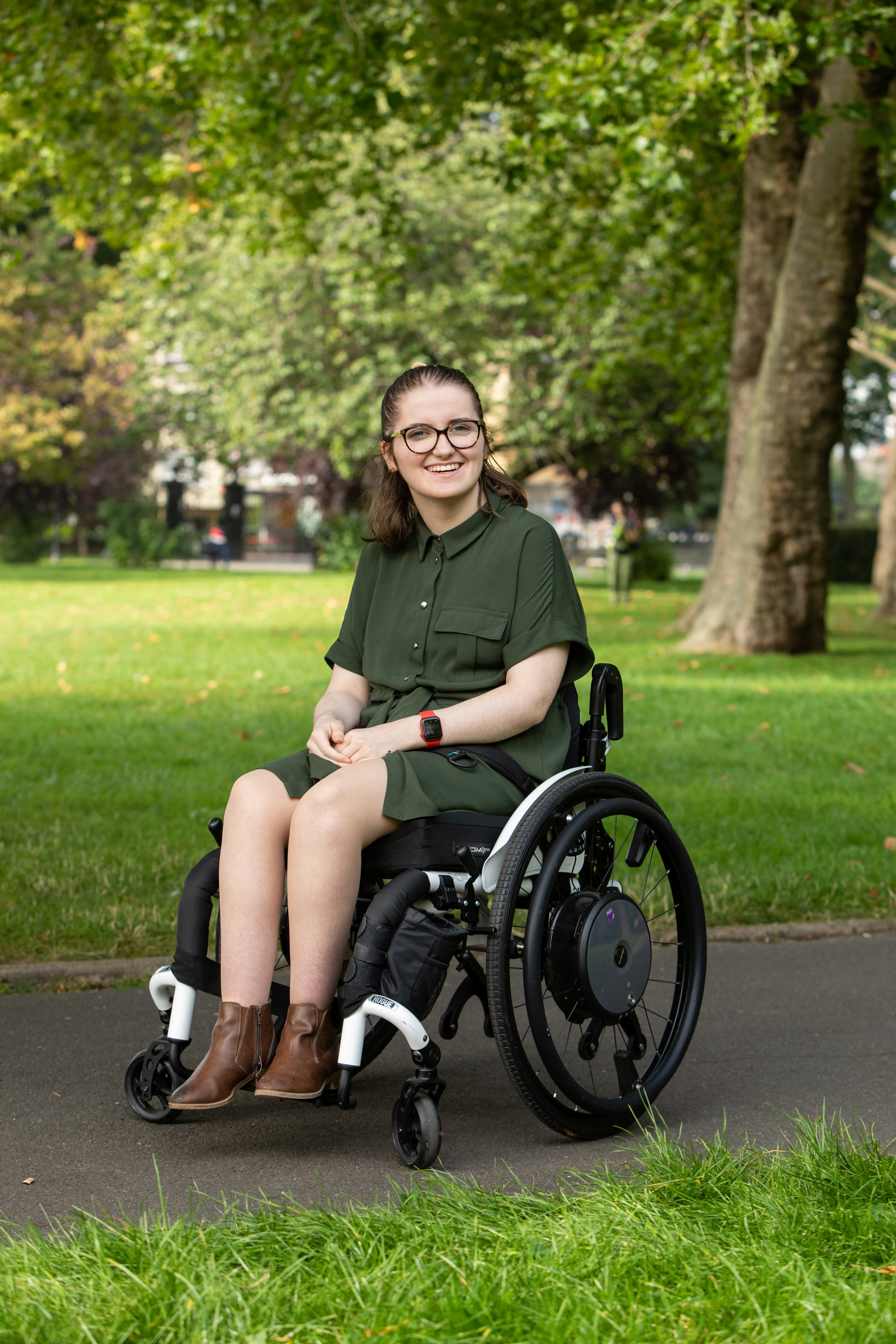 Penny sitting in wheelchair smiling, wearing glasses, khaki dress and brown ankle boots with grass and trees behind