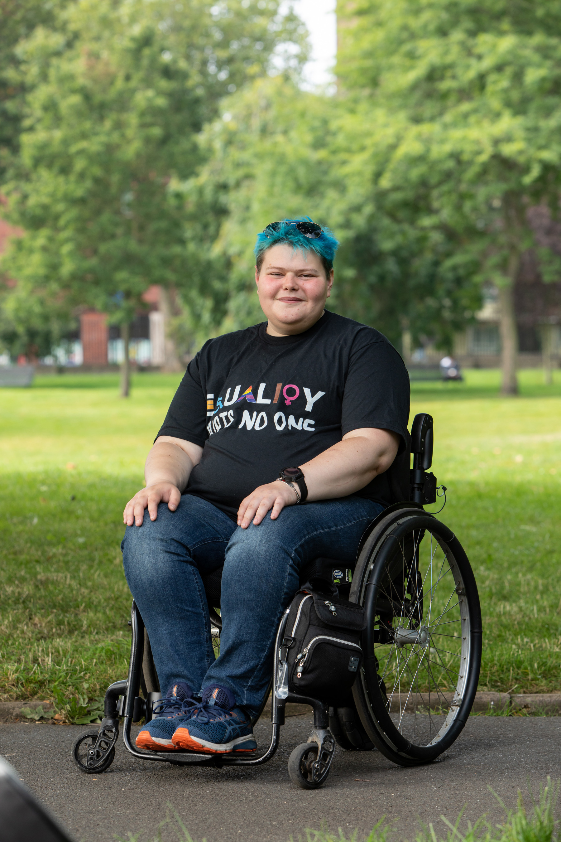 Eddie sitting in wheelchair smiling wearing Equality Hurts No One t-shirt, jeans and trainers with grass and trees behind