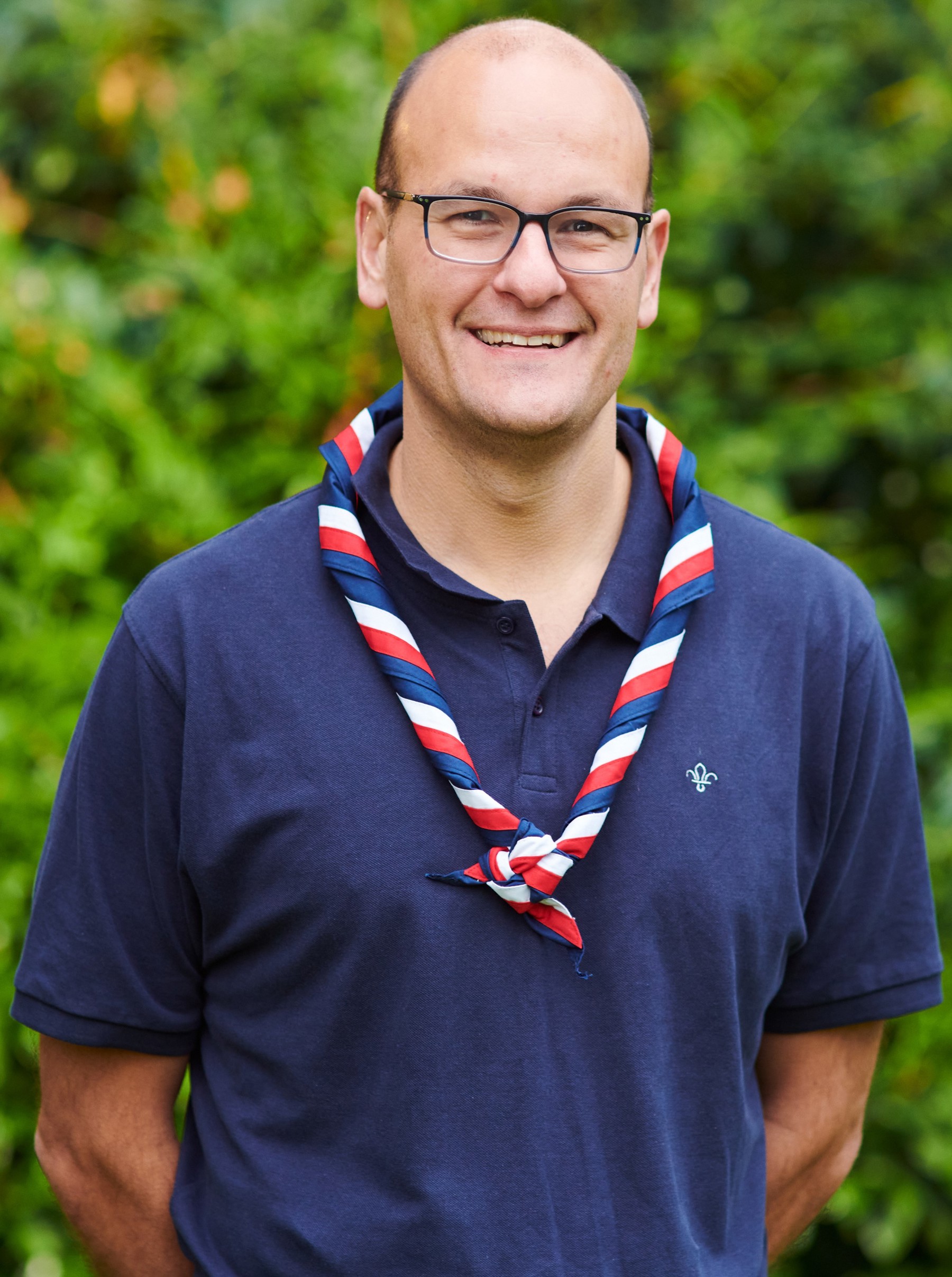 David Sandall smiling at the camera while wearing a navy Scouts polo shirt, glasses and stripy scarf