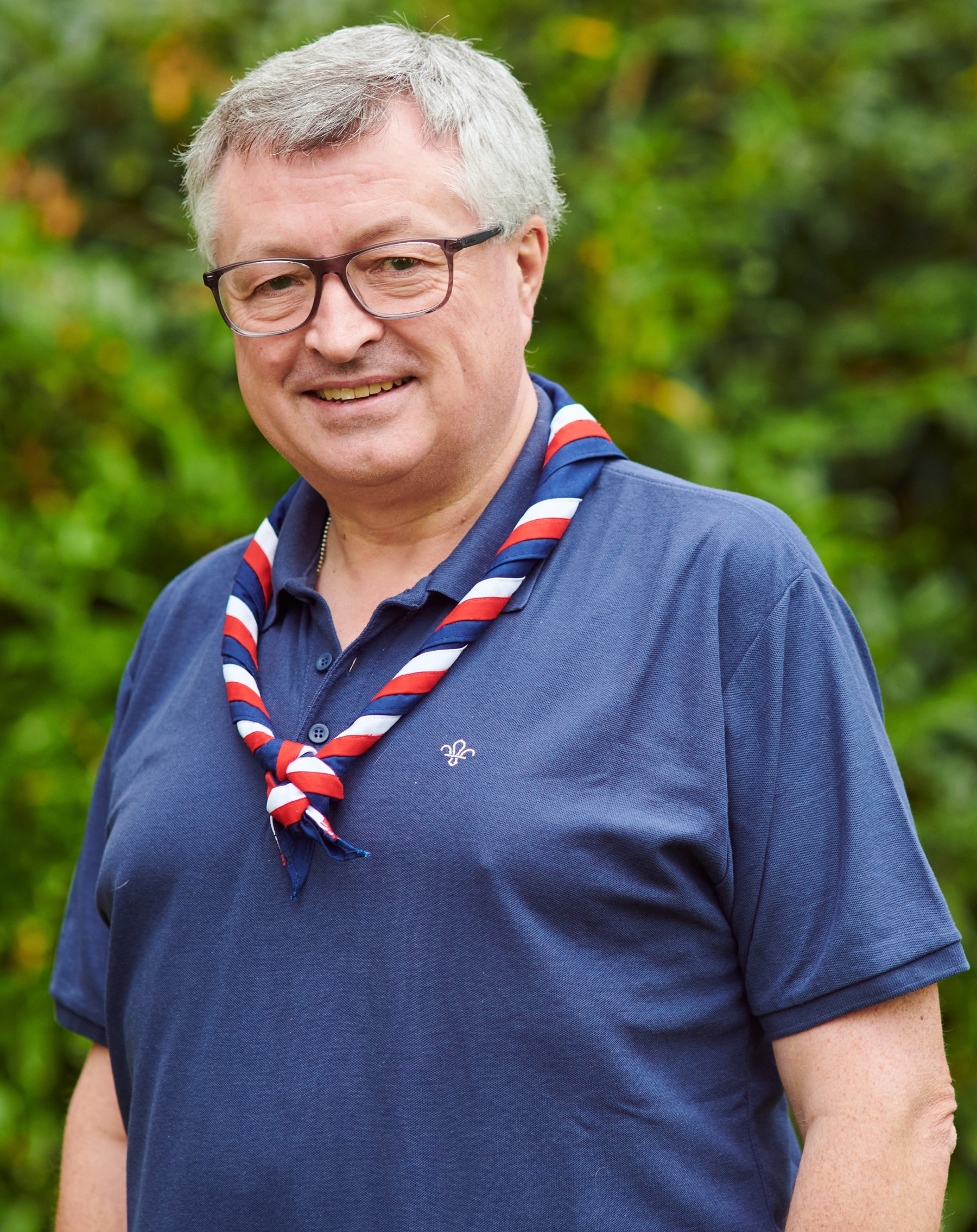 Graham Haddock smiling at the camera while wearing glasses, a navy Scouts polo shirt and stripy scarf