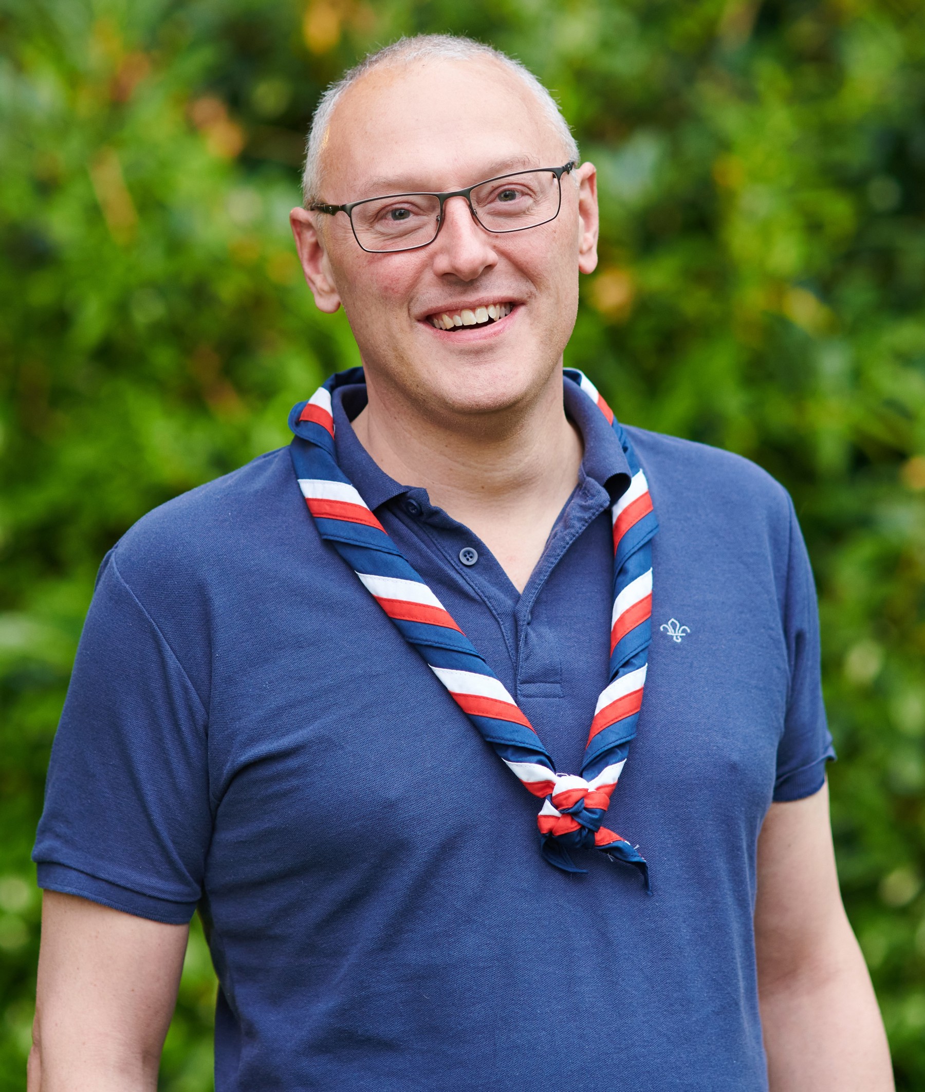 Nigel Ball smiling at the camera while wearing glasses, a navy Scouts polo shirt and stripy scarf