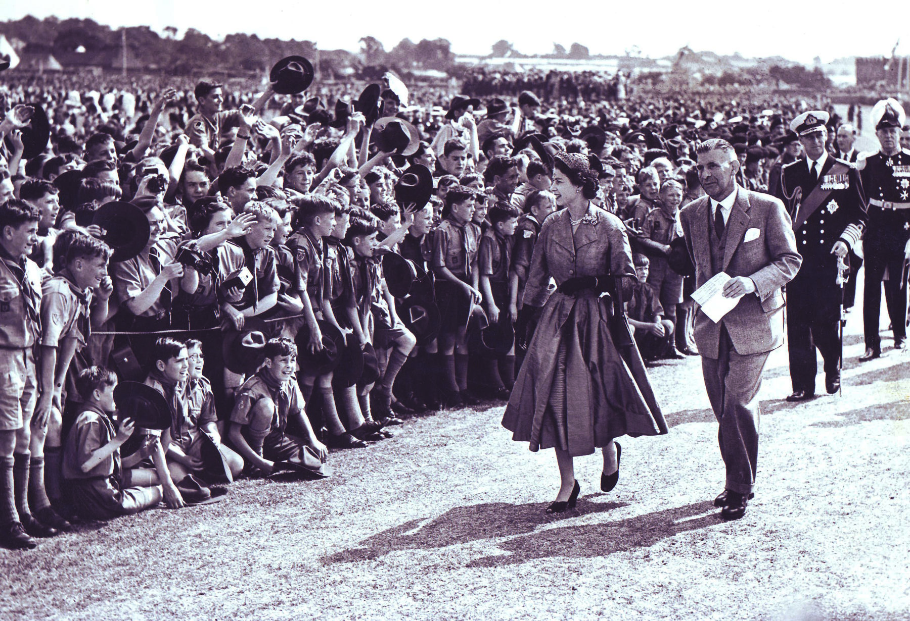 Sepia photo of the young Queen smiling walking past a cheering crowd of Scouts