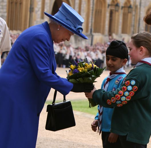 The queen wearing royal blue outfit being presented with a bouquet of flowers by a Beaver and a Scout