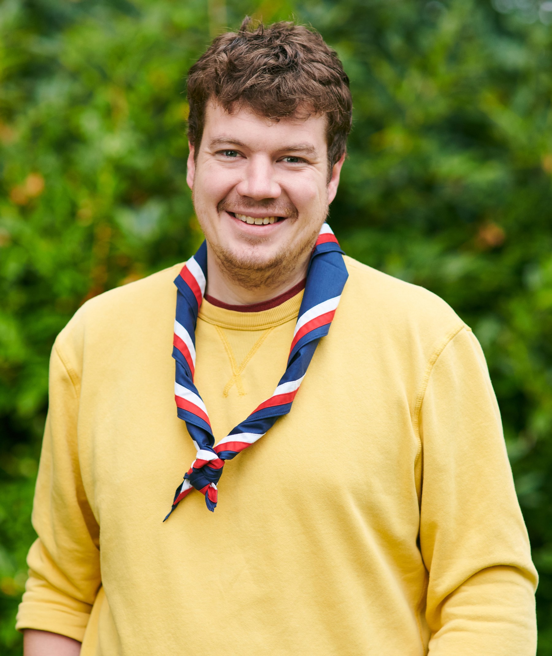 Jack Caine smiling at the camera while wearing a yellow sweater and stripy scarf