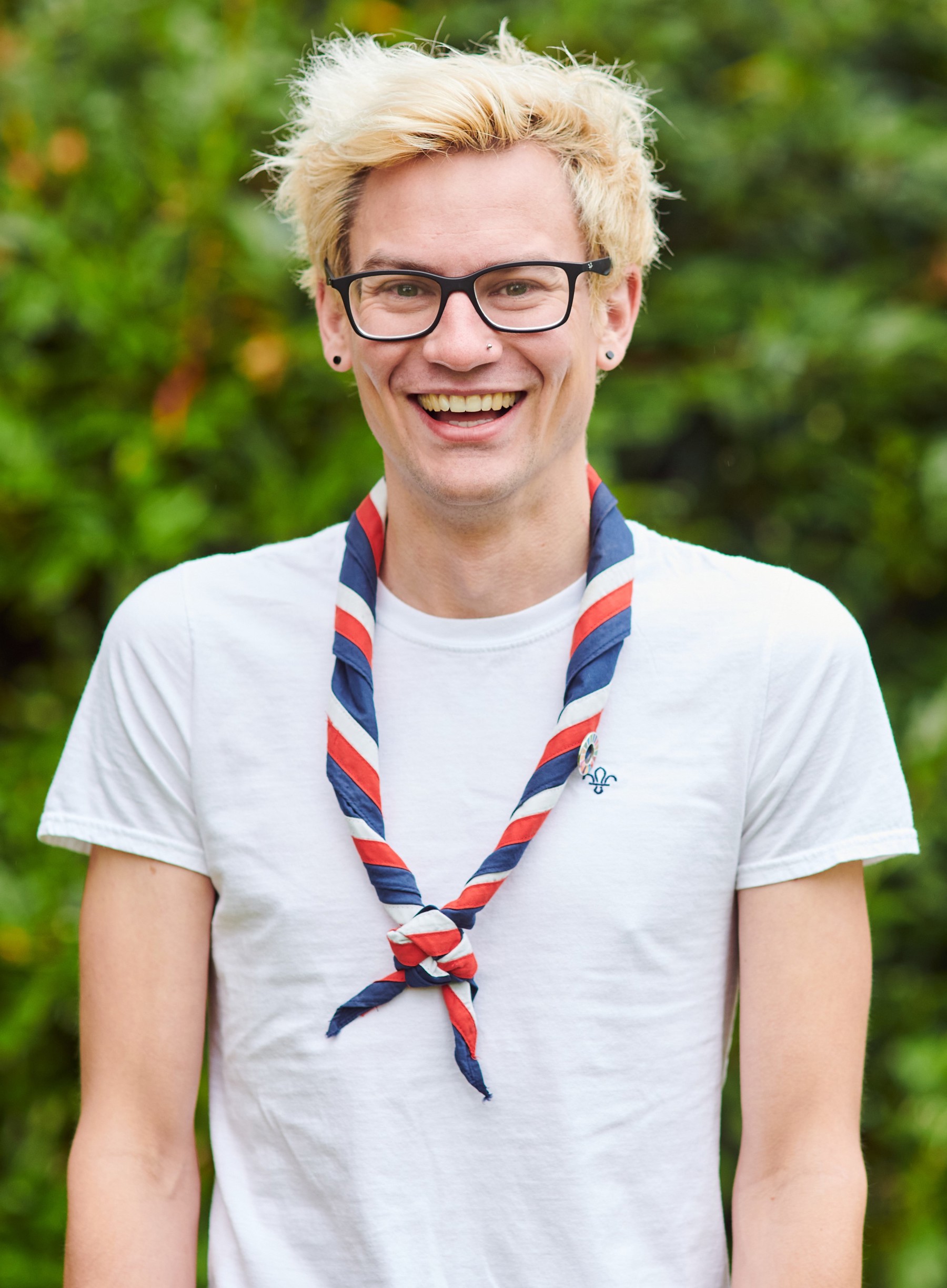 Tom Milson smiling at the camera while wearing a white Scouts t shirt and stripy scarf