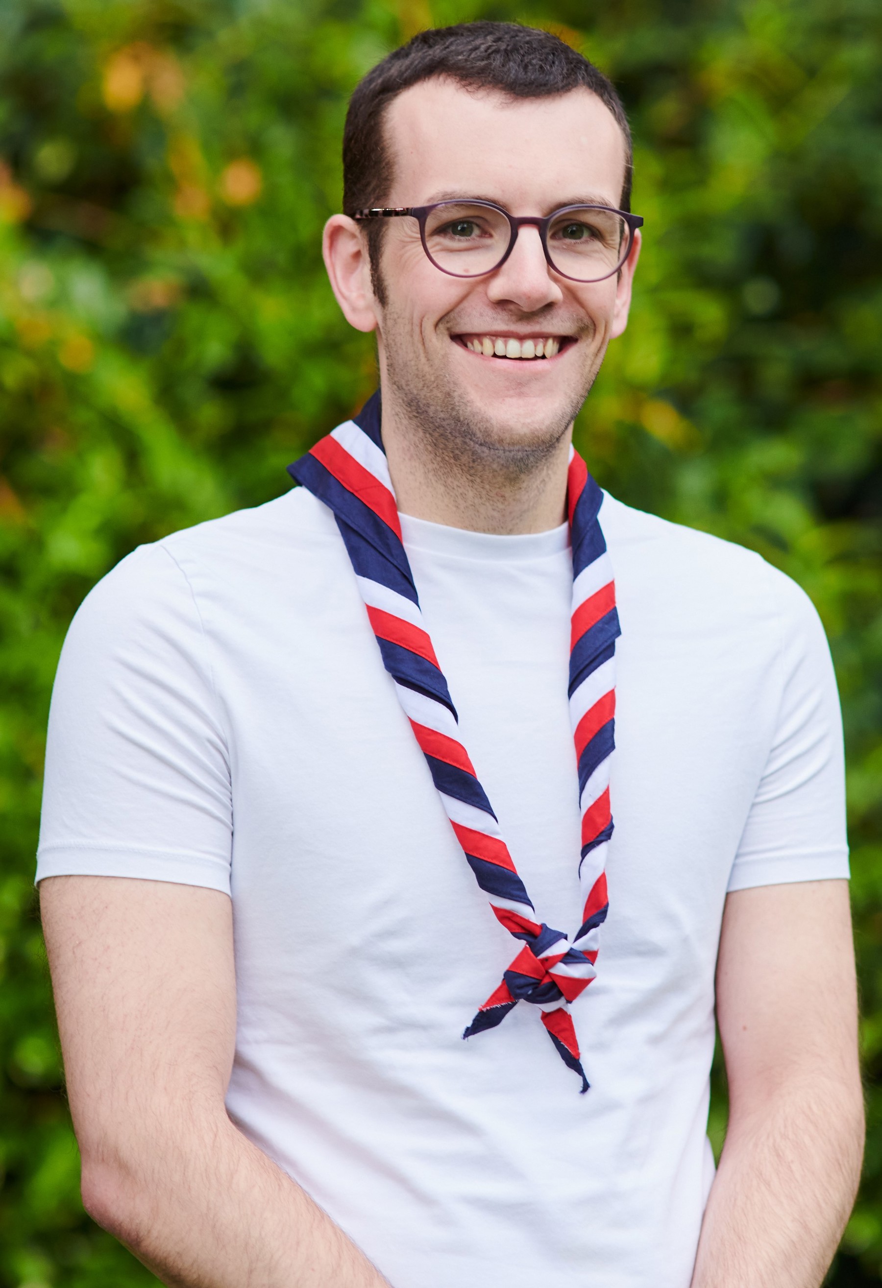 Lewis Dangerfield smiling at the camera while wearing a white t shirt and stripy scarf