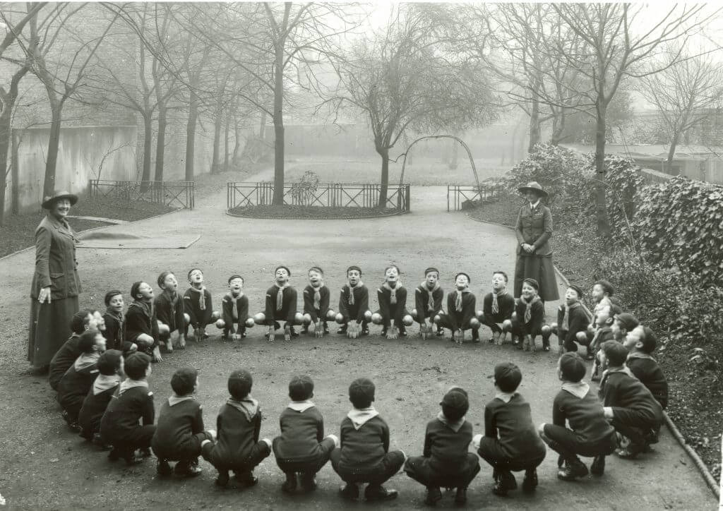 A group of Cubs wearing their uniform crouch in a circle while two leaders look over them