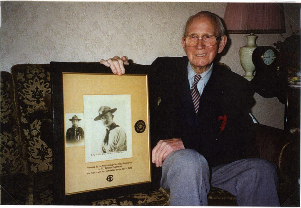 Image shows Arthur Shepherd in 1994 with a framed presentation panel showing a signed photo from Robert Baden-Powell, his coast watching badge and his Cornwell Badge