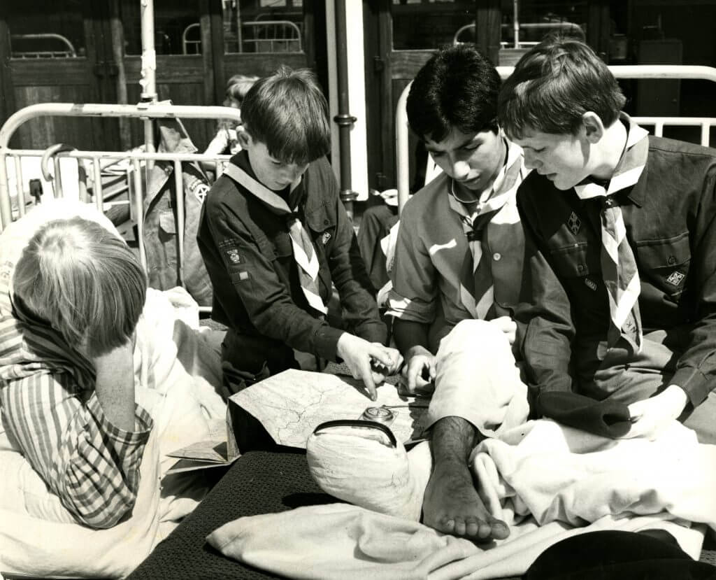 Black and white photo of a group of four Scouts leaning over a hospital bed, looking at a leg with a bandage on it