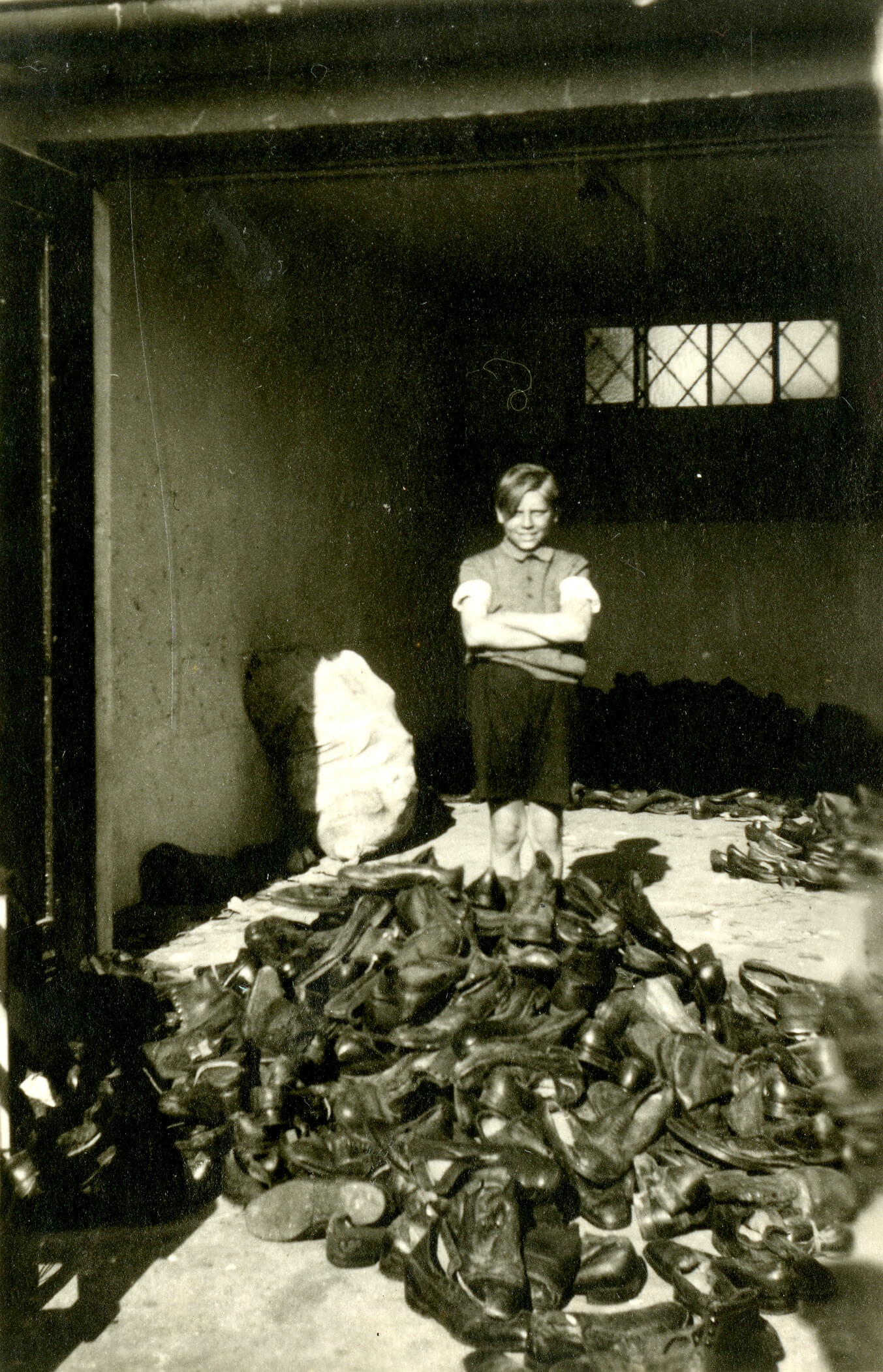 Black and white photo of a young person, arms crossed, standing behind a large pile of shoes