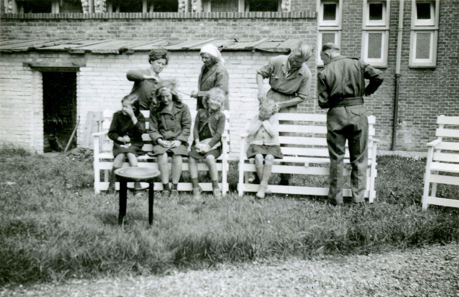 Black and white photo of four people sitting on benches having their hair checked for lice by four people standing behind them