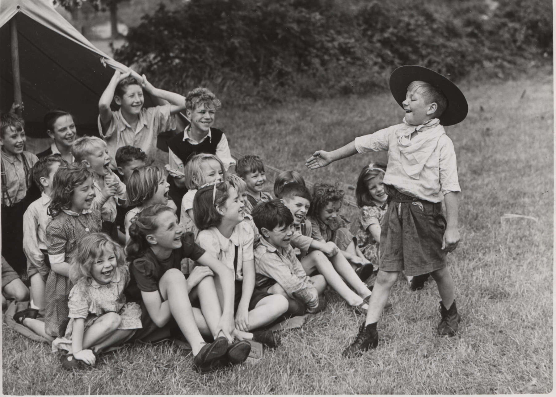 Black and white photo of a young person in Scouts uniform talking to a group of young people sitting down in front of a tent
