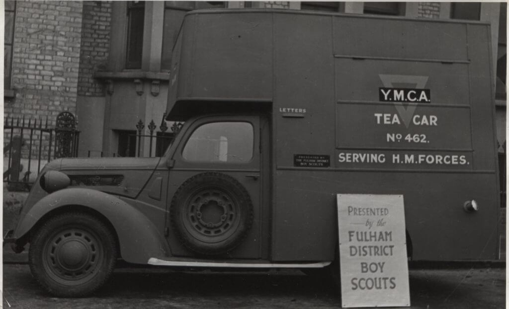 Black and white photo of the side of van, with YMCA Tea Car written on the side. A sign in front of it reads 'Presented by the Fulham District Boy Scouts'