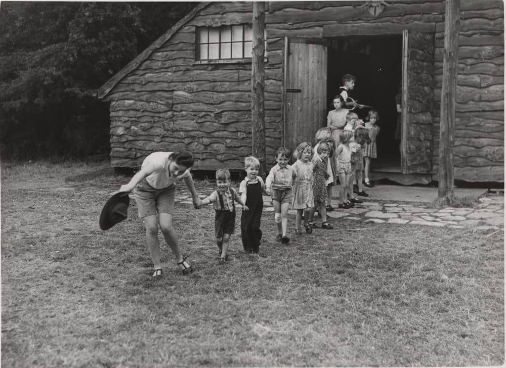 Black and white photo of a group of young people holding hands as an adult leads them in a line out of a house