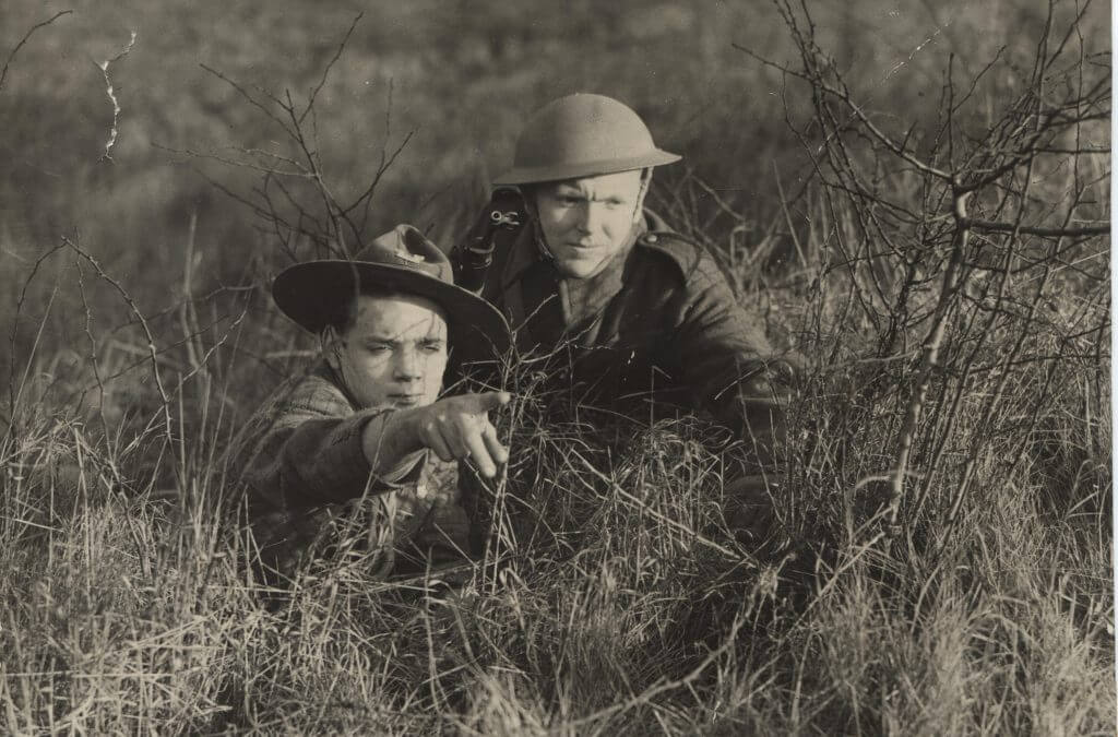 Black and white photo of a Scout and an adult laying in grass. The Scout is pointing ahead, showing something to the adult.