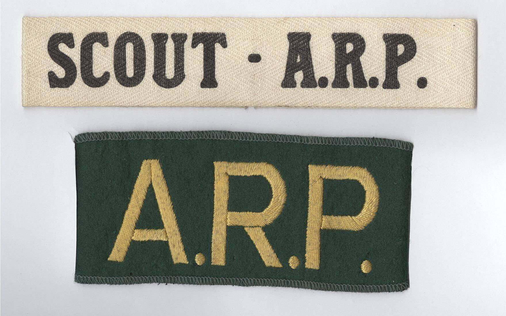 The armbands. The top one is cream with 'SCOUT - A.R.P.' written on it. The bottom one is green and wider, with 'A.R.P.' written in yellow stitching.