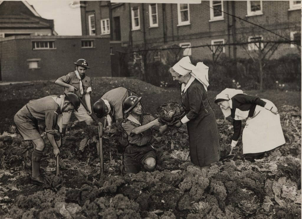 Black and white photo of a group of Scouts gardening in a muddy field with two nurses