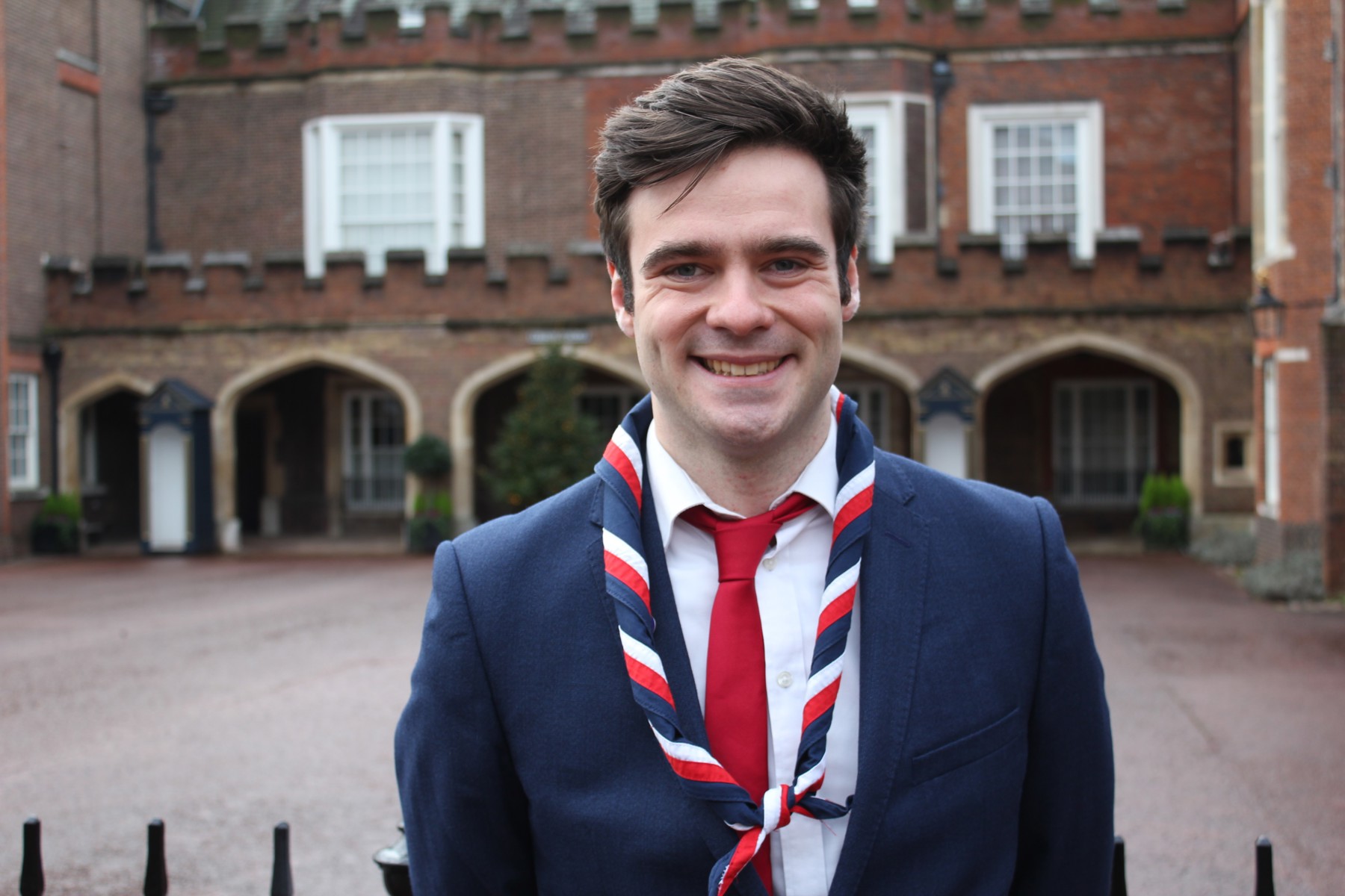 Jack, wearing a white shirt, navy jacket, red tie and stripy scarf, smiles at the camera