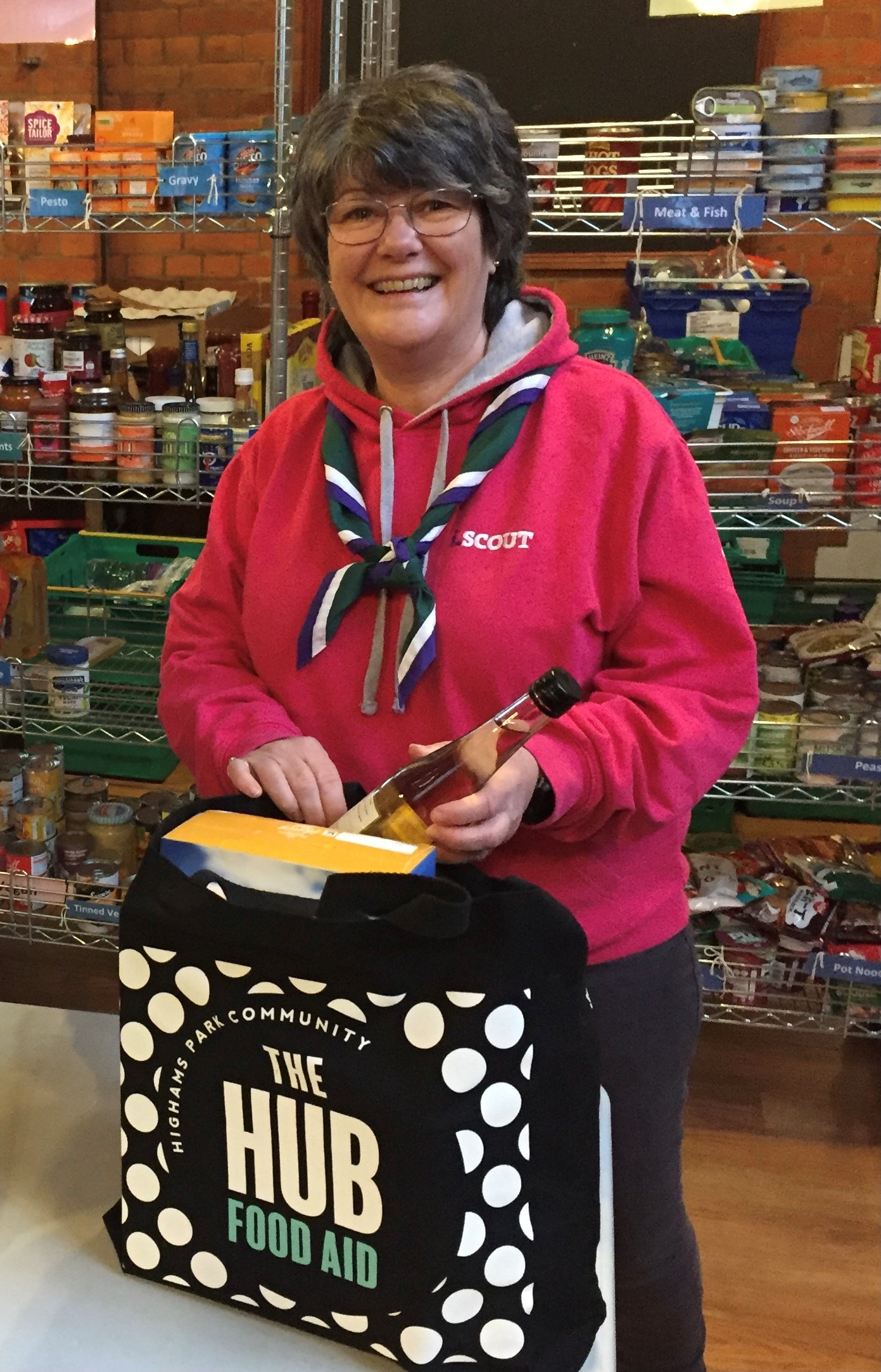 Tracey, wearing a red Scouts hoodie, places a bottle into a bag while smiling at the camera