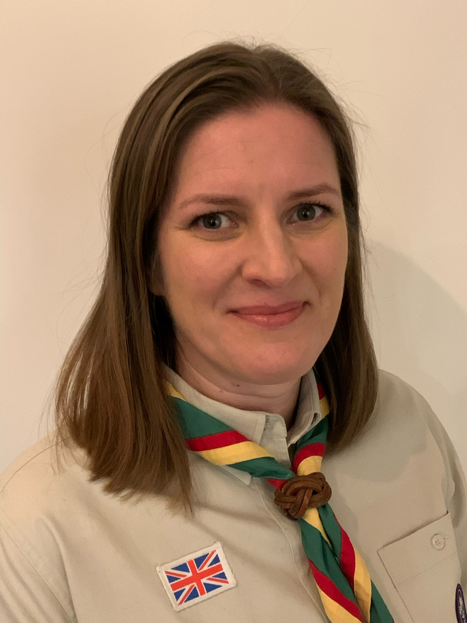 Sarah, wearing a beige Scouts shirt and stripy scarf, smiles at the camera