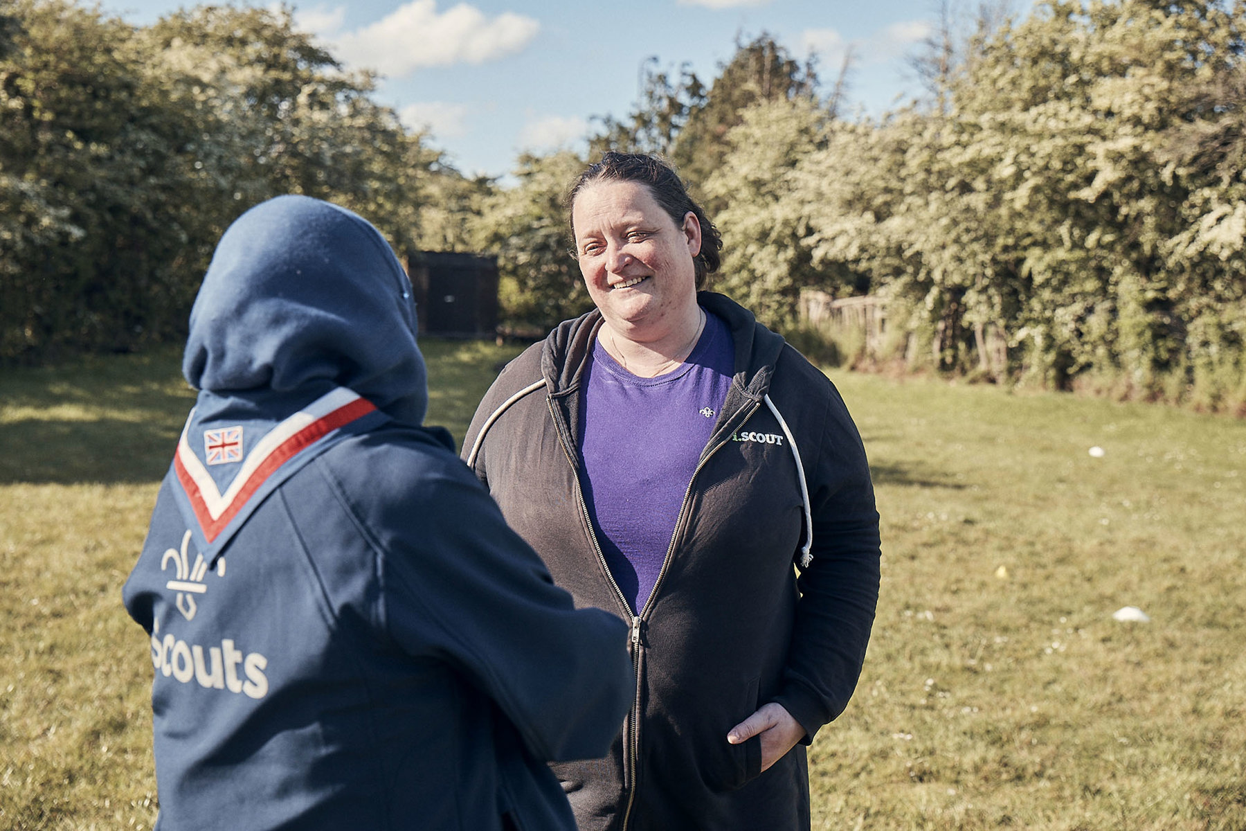 Two female volunteers chatting amiably outdoors with green space behind, both in Scouts uniforms, one with a Muslim headscarf