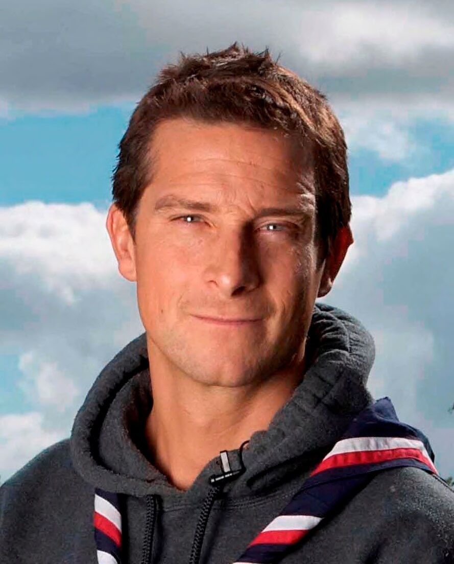 Bear Grylls looks at the camera, smiling slightly, while wearing a dark grey hoodie and red, white and blue scarf.