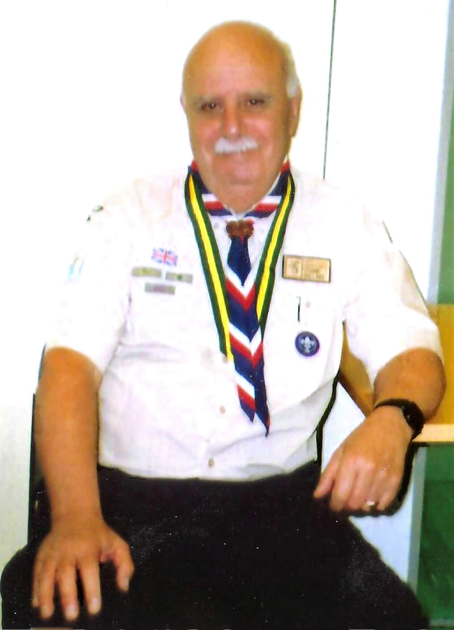 Stuart, wearing a white short-sleeved shirt, red, white and blue scarf and green and yellow medal, smiles at the camera while sitting in a chair.