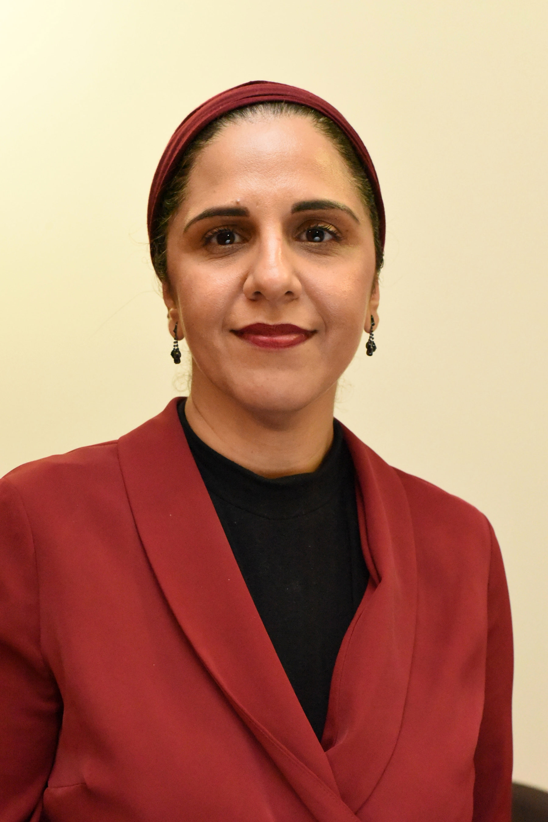 Fozia Irfan smiling at the camera while wearing a red suit jacket with matching red hijab, and black jumper and earrings