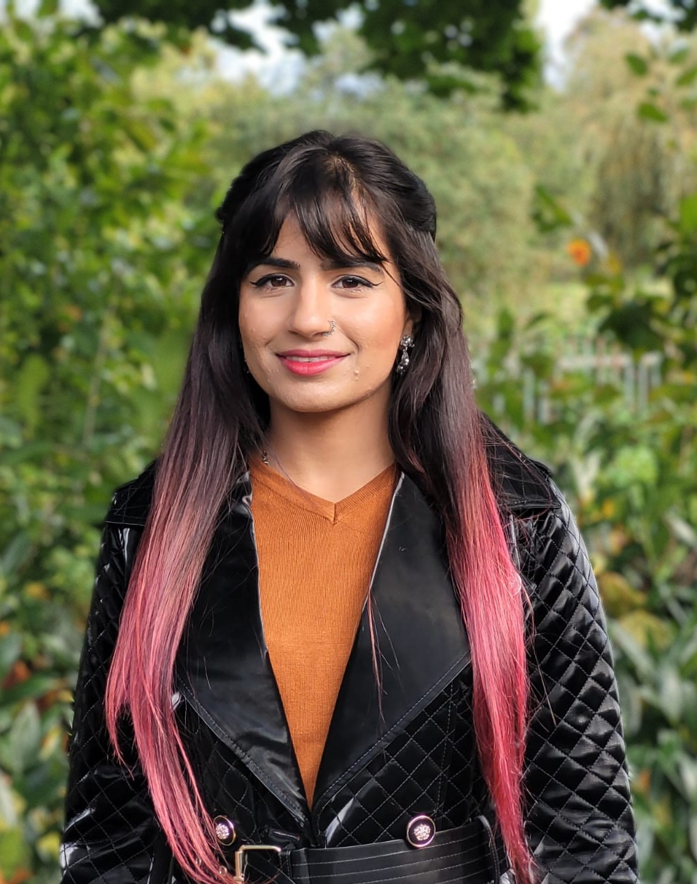 Rehneesa Inez smiles at the camera in front of a background of trees, wearing black jacket and orange jumper underneath