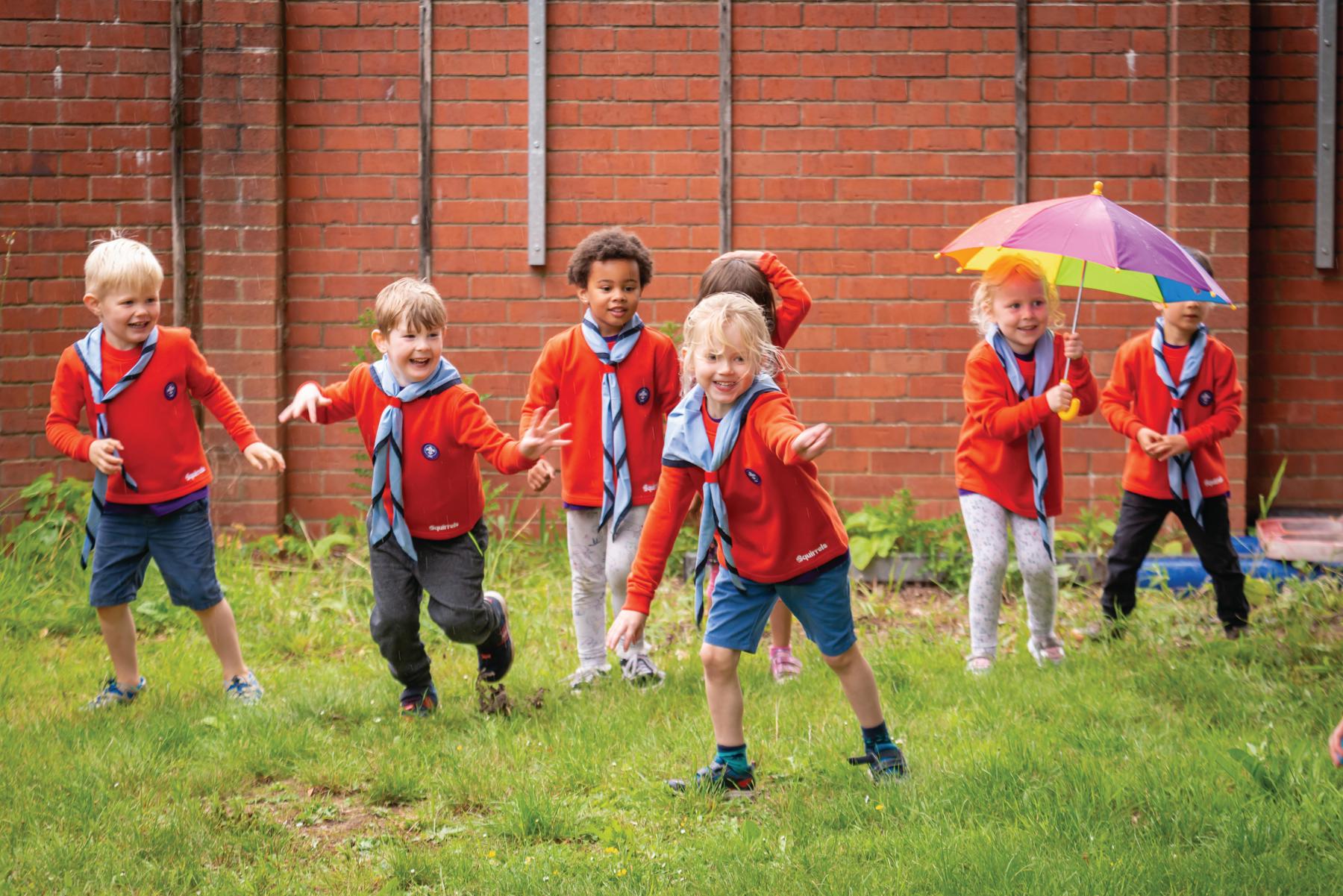 A group of Squirrels are playing on a patch of grass in front of a wall. They are playing a running came and all looking over to the right. They are all wearing red jumpers with blue neckers and have big smiles. There are four boys and three girls and they're all having lots of fun. One girl is even holding a rainbow umbrella!