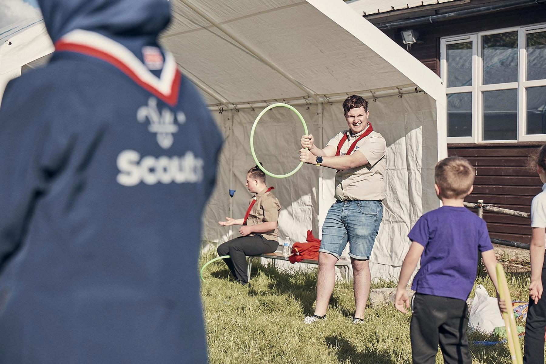 A Scout holds a hoop in the air with a young boy in a purple t shirt looks ready to throw something through it. They are outside their meeting place on grass and underneath a marquee.