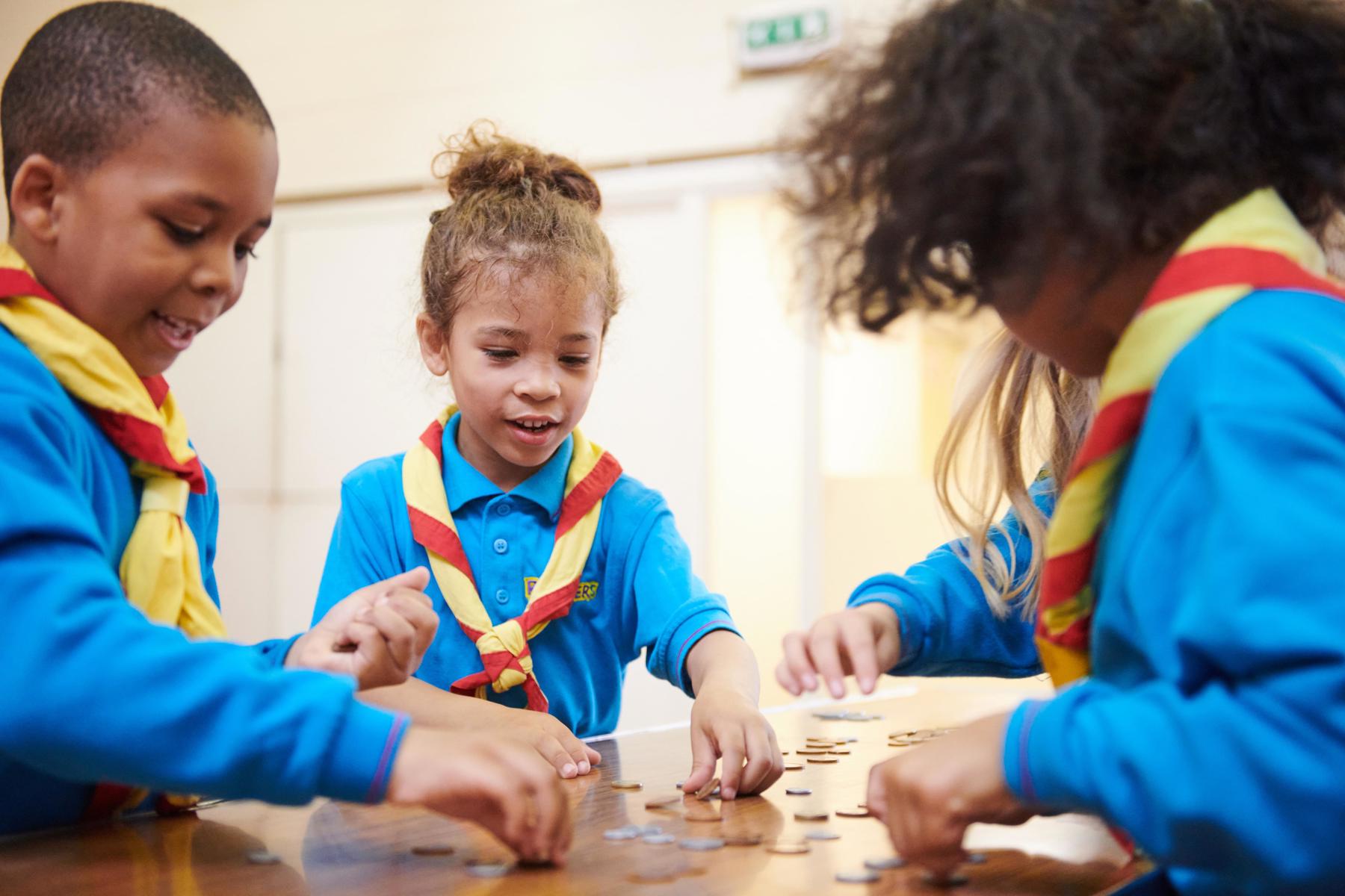 A group of Beavers are stood round a table playing with pretend money and are reaching for the coins.