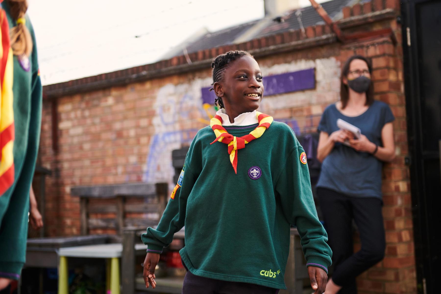 A young cub in a playground. He is playing a game and smiling, with his Cubs jumper on. He has badges down both arms and a yellow and red necker.