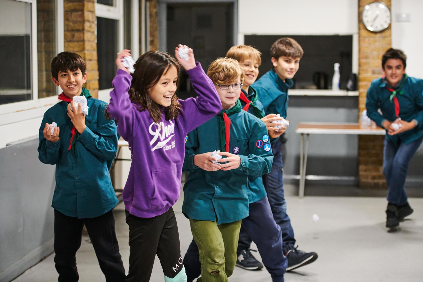 A group of Scouts are inside their meeting place. There are six boys and a girl. They are playing a throwing game, running around and holding balls of paper to throw. The girl at the front looks very excited, as she is holding balls of paper in both hands and has both arms in the air, ready to throw them. She is wearing a purple hoodie.